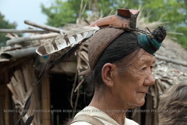 L’anziano Nyishi porta ancora il tipico copricapo a forma di casco di bambù sormontato dal becco del bucero con 3 piume di pavone o bucero dietro, portando i capelli annodati sulla fronte e fermati con degli spilloni di ottone